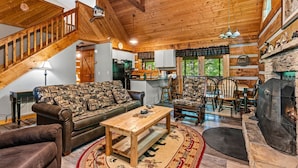 Ein gemütliches Blockhaus-Interieur mit einer Holztreppe, einem Wohnbereich mit Sofa und Couchtisch, einem Essbereich mit Stühlen und einer Küche mit Kühlschrank und Herd.
