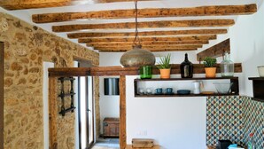 Uma cozinha rústica com vigas de madeira aparente, paredes de pedra e uma estante com diversos utensílios de cozinha.