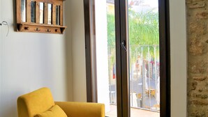 Um quarto com uma poltrona amarela, um banquinho de madeira, uma prateleira de madeira com ganchos e uma janela grande com vista para o exterior.