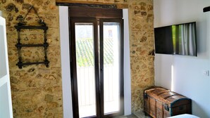 Um quarto com paredes de pedra, uma porta de madeira, uma televisão fixada na parede e um baú de madeira.