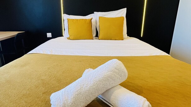 Een net opgemaakt bed met wit en geel beddengoed, een opgerolde witte handdoek en een zwart hoofdeinde met verlichte lijnen.