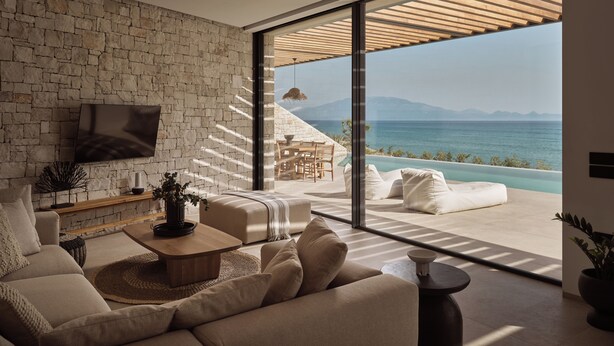 Un salón moderno con una pared de piedra, un televisor de pantalla plana, un sofá, una mesa de centro y vistas al mar a través de puertas de cristal amplias.