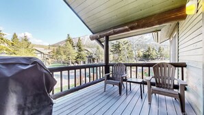Een houten terras met twee Adirondack-stoelen en een barbecue, met uitzicht op een woonwijk met huizen en bomen.
