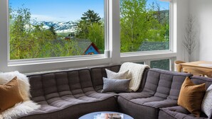 Un salon moderne avec un canapé d’angle, une petite table et de grandes fenêtres donnant sur un paysage de montagne et de forêts.