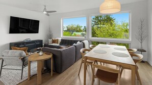 Un salon moderne comprenant un canapé, une table à manger avec des chaises, un ventilateur de plafond et de grandes fenêtres donnant sur un paysage verdoyant.