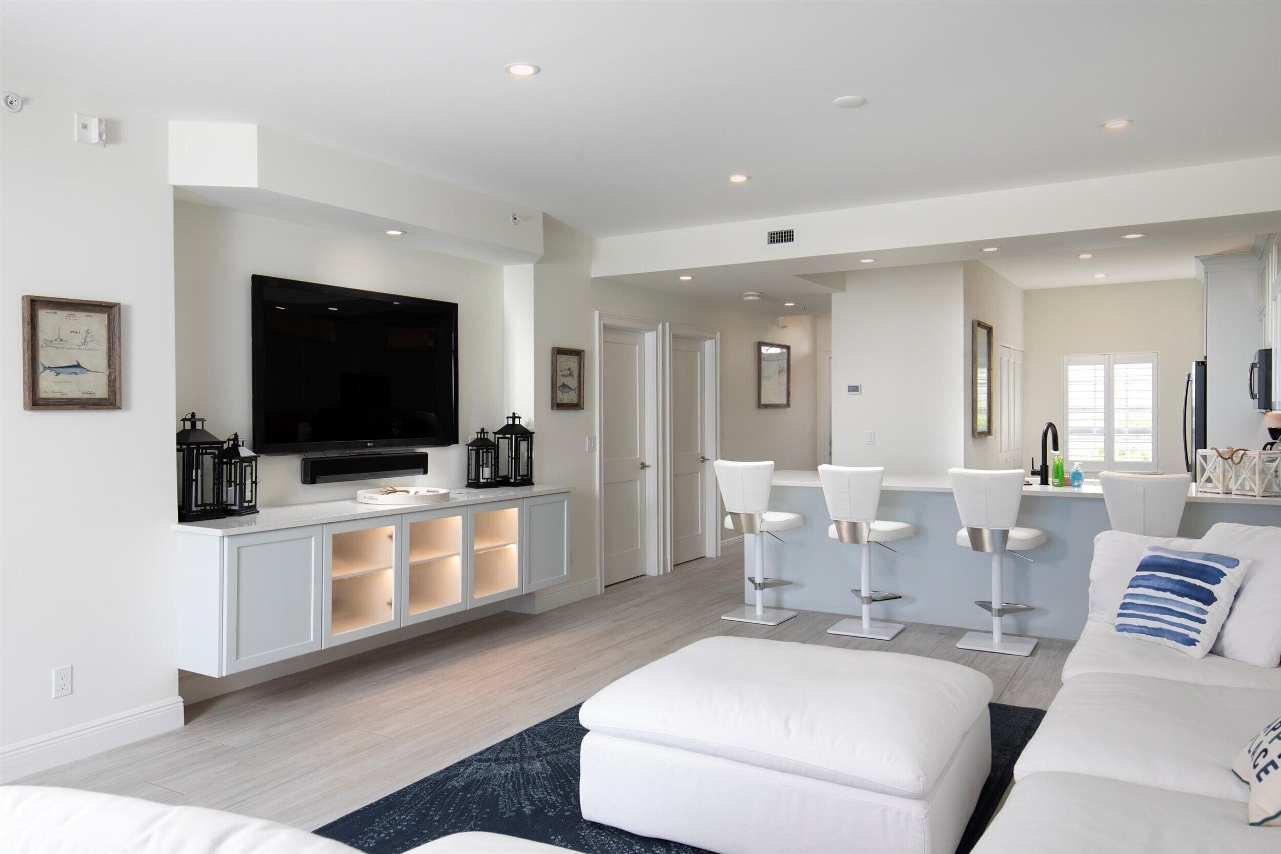 A modern living room with a large flat-screen TV, white sofas, a coffee table, and a bar area with stools.