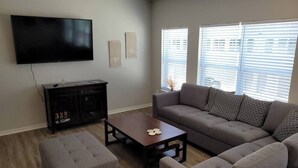 Un salon doté d’un téléviseur à écran plat, d’un meuble de rangement en bois foncé, d’un canapé gris agrémenté de coussins à motifs, d’un pouf assorti et d’une table basse.
