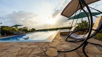 A poolside area with lounge chairs and umbrellas, a stone-tiled deck, and a clear view of the sky.