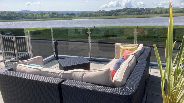 En balkon med fletmøbler, gelænder af glas og udsigt over en flod og bakker.