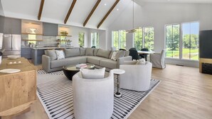 A spacious living area with a large L-shaped sofa, a coffee table, and a dining table. There is a kitchen area with modern appliances and a large window offering a view of the outdoors. - 4 BR Luxury Home w Private Pool | New Renovation (Isle of Palms)
