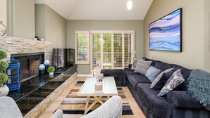 A living room with a stone fireplace, a black sofa, a coffee table, and a large painting on the wall. - Gated Gainey Ranch condo, garage, heated pool, walk to restaurants, shopping! (Scottsdale)