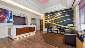 A modern hotel lobby with a reception desk, a large abstract wall mural, and a seating area with leather sofas. - Lidos Hotel (London)
