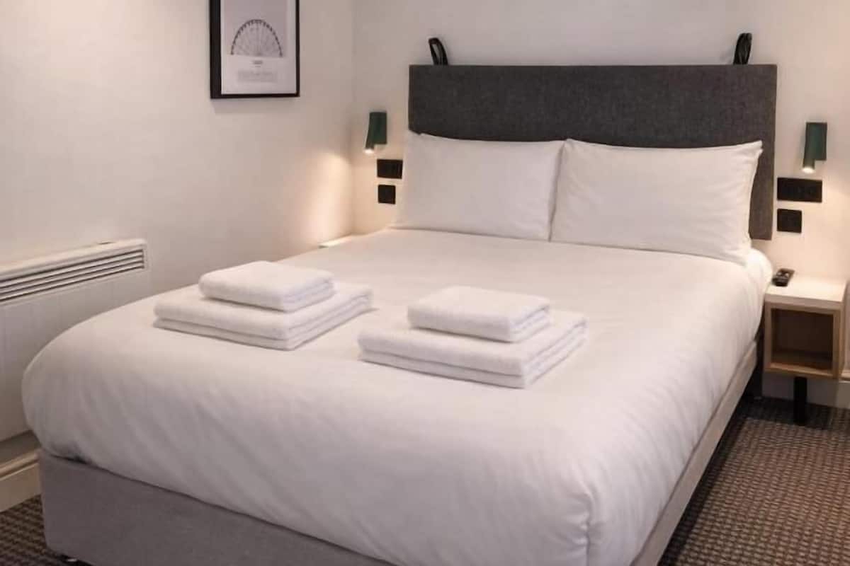 Un lit bien fait, avec du linge de lit blanc et des serviettes pliées, une tête de lit rembourrée de couleur grise et deux tables de chevet avec des lampes fixées au mur.