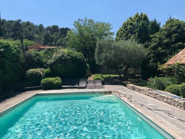 Relax by the serene pool surrounded by lush greenery and mature trees, with lounge chairs and a stone wall.