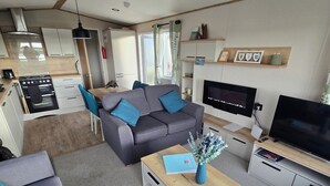 Un espacio habitable compacto con kitchenette, un sofá gris con cojines azules, un televisor de pantalla plana y una pequeña mesa de centro de madera.