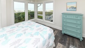 Un dormitorio con una cama grande, un aparador y vista a la playa a través de amplias ventanas.