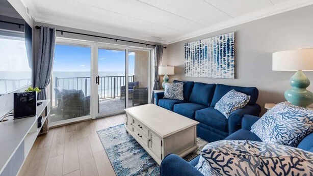 Een moderne woonkamer met een blauwe zithoek, een witte salontafel en een groot raam met uitzicht op zee.