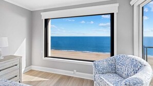 Een kamer met een groot raam met uitzicht op een strand, een witte dressoir en een gestreept fauteuil.