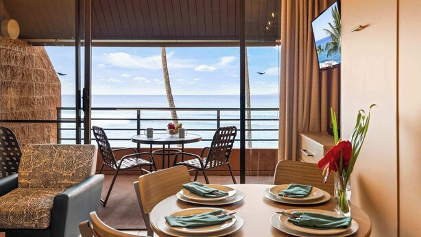 A dining area with a table set for four, overlooking a beach with palm trees and the ocean. - Kuleana Club #619 Oceanfront - 1 bd & 1 ba- Gated (Lahaina)