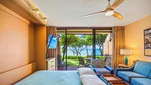 A hotel room with a bed, a seating area with a view of the ocean, a ceiling fan, and a TV mounted on the wall.