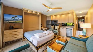 A compact hotel room with a bed, a desk, a kitchenette, and a ceiling fan.