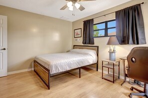 A bedroom with a bed, a desk, a chair, a ceiling fan, and a window with curtains.