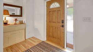 Une porte en bois avec un panneau de verre, un mur blanc, un plancher en bois, un meuble beige, un miroir, une lampe et une petite plante.