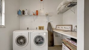 A laundry room with two white Bosch washing machines, a shelf with cleaning supplies, and a 'Bless This Home' sign. - Sunny Pool Retreat, Pet-Friendly Near Beaches (Naples)