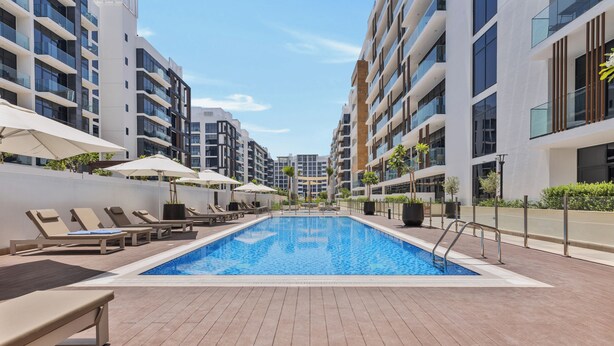 Un espace piscine moderne, doté de chaises longues et de parasols, entouré d’immeubles de grande hauteur.