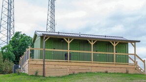 A small, single-story building with a green exterior and a wooden porch. It features a metal roof and is situated on a grassy hill with trees in the background. - Safari Tent A at Eden Reserve - Near Ark (Williamstown)