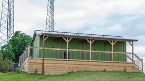 Ein kleines, eingeschossiges Gebäude mit grüner Fassade und einer Holzveranda. Es hat ein Metall-Dach und steht auf einem grasbewachsen Hügel mit Bäumen im Hintergrund.