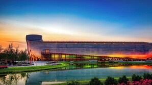A large, modern building with a unique curved roof, surrounded by a reflective water body and landscaped gardens.