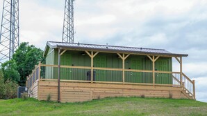 A small, single-story building with a green exterior and a wooden porch. It features a metal roof and is situated on a grassy hill with trees in the background. - Safari Tent D at Eden Reserve - Near Ark (Williamstown)