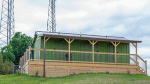 A small, single-story building with a green exterior and a wooden porch. It features a metal roof and is situated on a grassy hill with trees in the background.