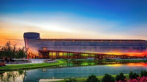 Un edificio grande y moderno con un techo curvo distintivo, rodeado por una superficie acuática reflectante y jardines ajardinados.