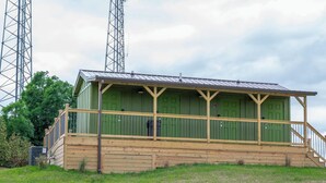 Ein kleines, eingeschossiges Gebäude mit grüner Fassade und einer Holzveranda. Es hat ein Metall-Dach und steht auf einem grasbewachsen Hügel mit Bäumen im Hintergrund.