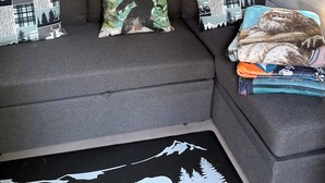 A grey sectional sofa with three patterned throw pillows and a blanket, against a wooden wall with framed artwork and wall decals. - Property of Big Foot Roscoe, NY (Roscoe)