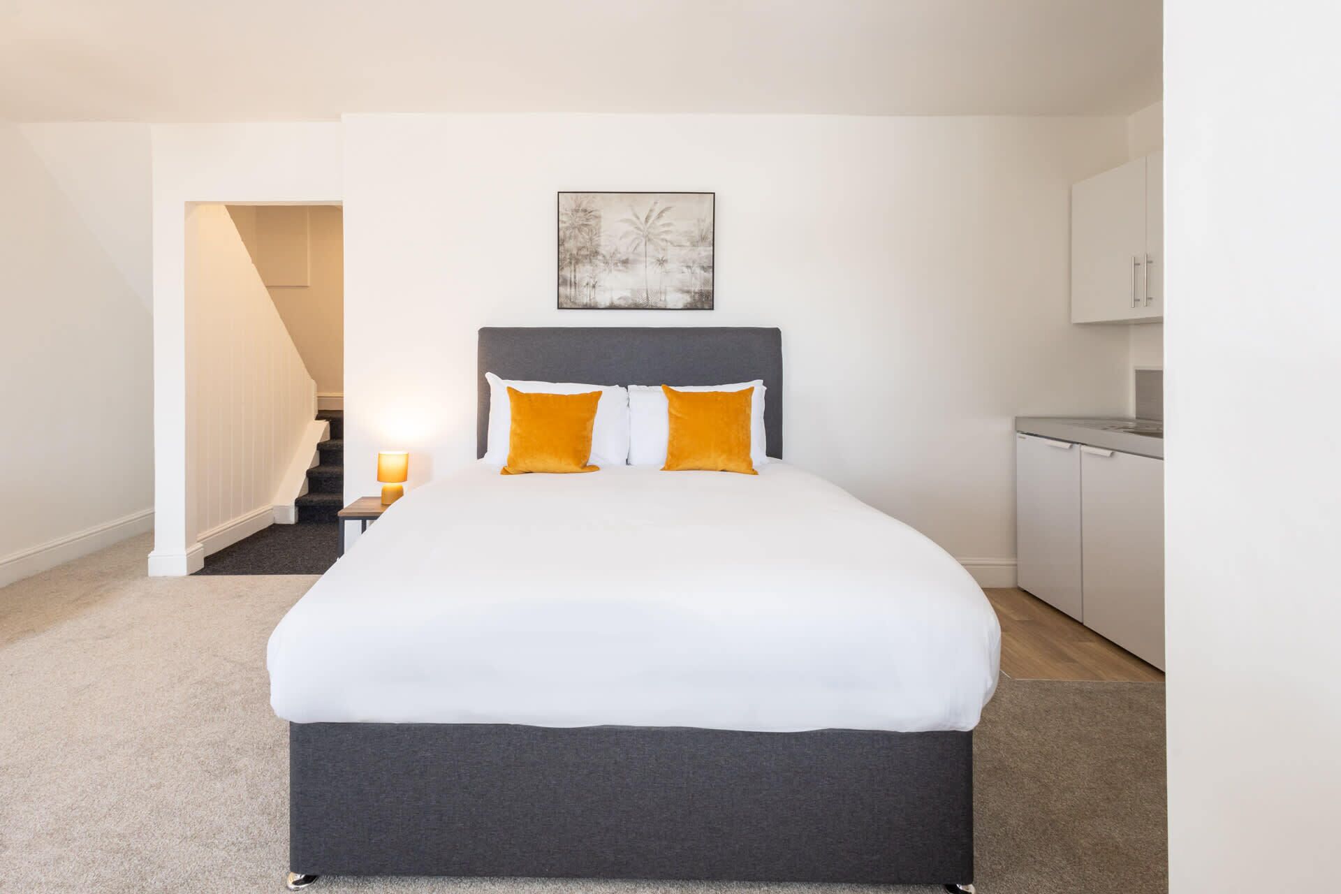 Una camera da letto con un letto, un comodino, una lampada, un quadro e un angolo cottura.