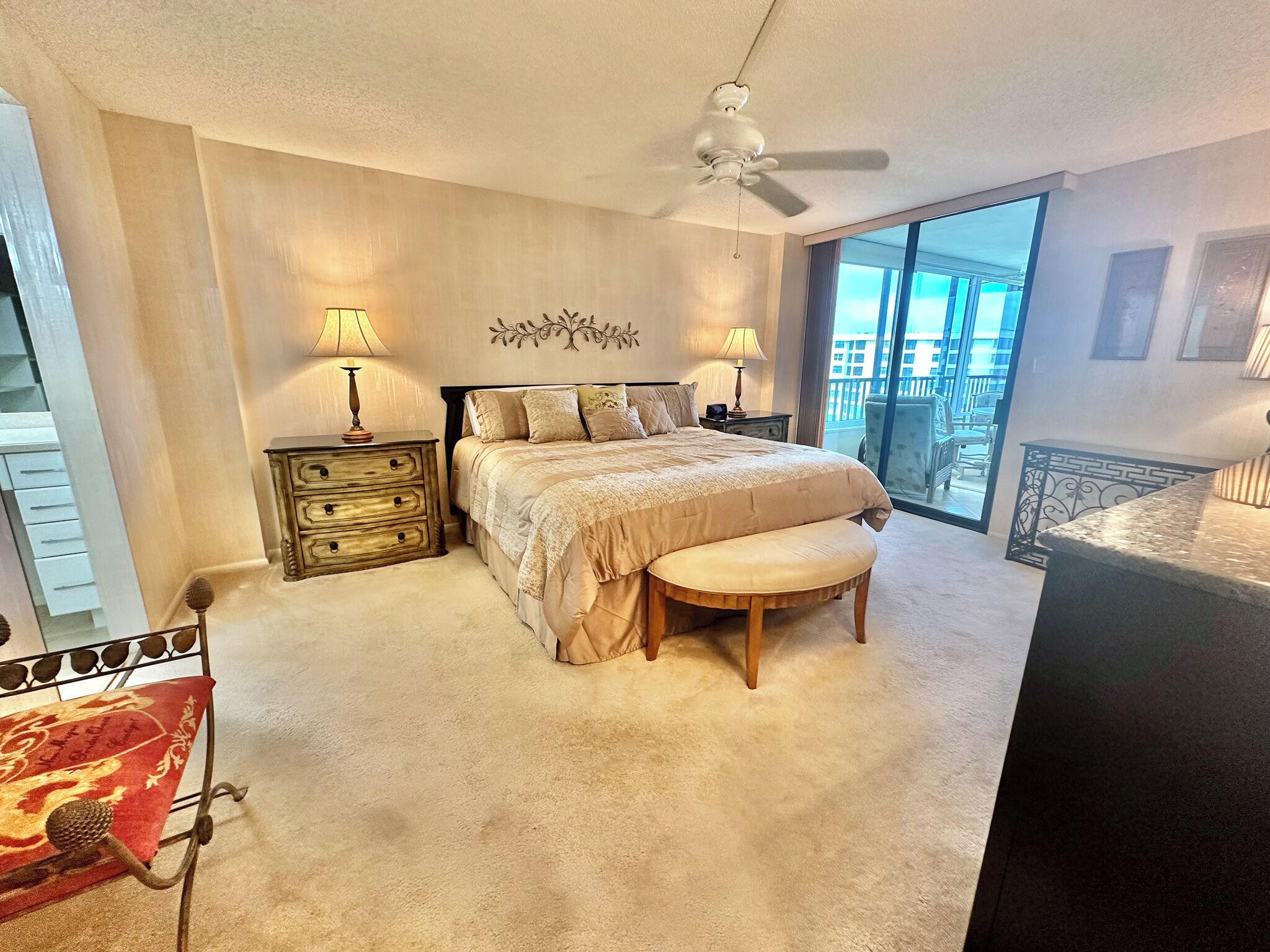 A bedroom with a bed, bedside tables, a bench, a ceiling fan, and a large window with a view.