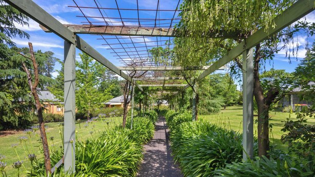 Un sentier de jardin agrémenté d’une pergola, entouré d’une végétation luxuriante et surplombé d’un ciel dégagé.