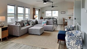 Ein geräumiges Wohnzimmer mit einer Eckcouch, einem gemusterten Sessel und einem Essbereich im Hintergrund.