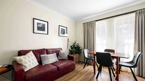 A living room with a red leather sofa, a dining table with chairs, a potted plant, and two framed pictures on the wall. - Melview Greens - Two-Bedroom Apartment (Orange)