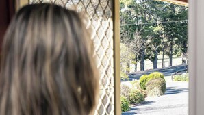 A person with blonde hair standing at a doorway with a lattice design, looking out onto a paved area with trees and bushes. - Melview Greens - Two-Bedroom Apartment (Orange)