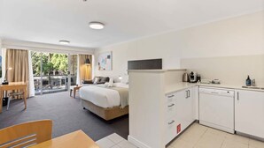A hotel room with a bed, a desk, a dining table, a kitchenette, and a balcony with a view.