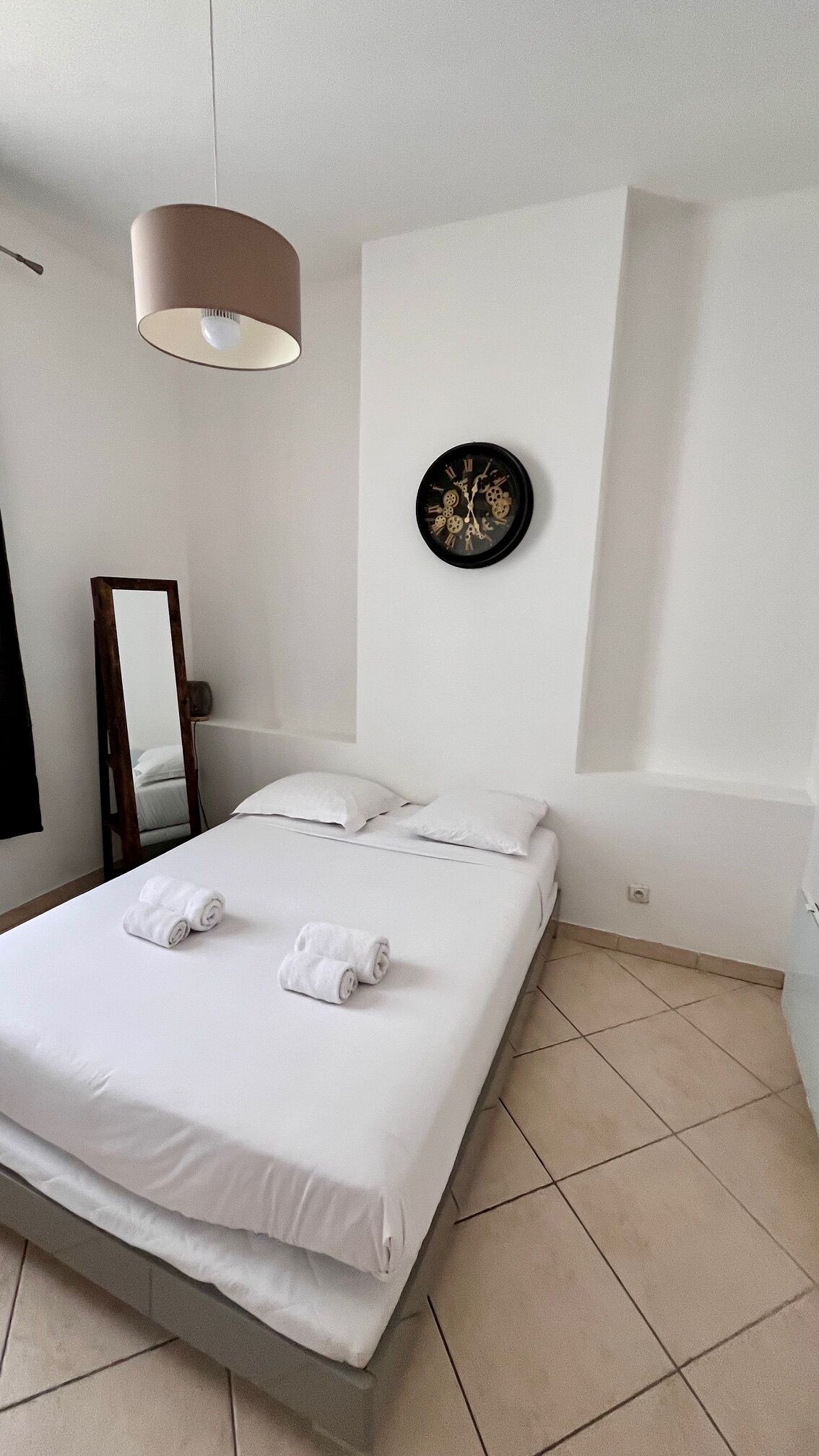 A hotel room with a bed, a wall clock, a mirror, and a hanging light fixture.