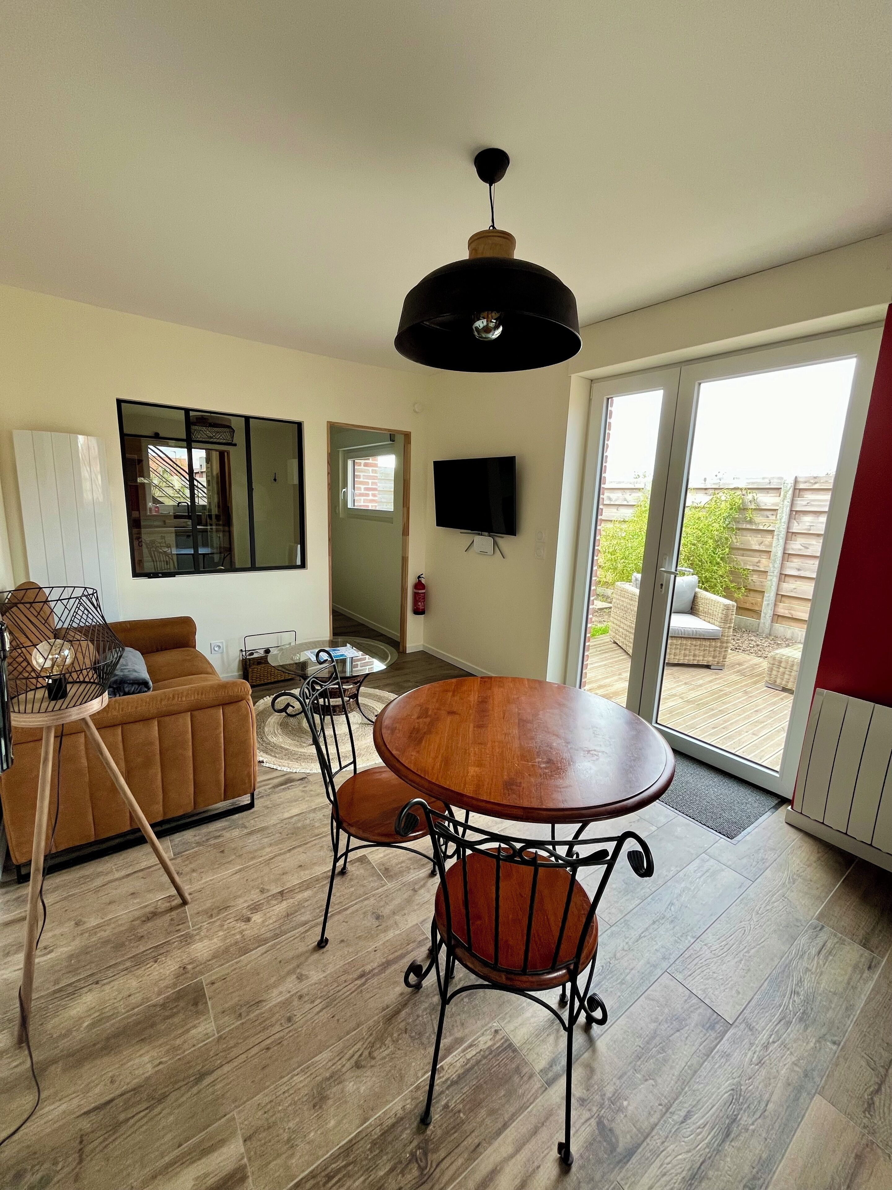 A modern living room with a sofa, a dining table with chairs, a TV mounted on the wall, and a sliding glass door leading to an outdoor area.