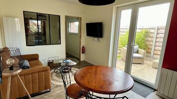 A modern living room with a sofa, a dining table with chairs, a TV mounted on the wall, and a sliding glass door leading to an outdoor area.