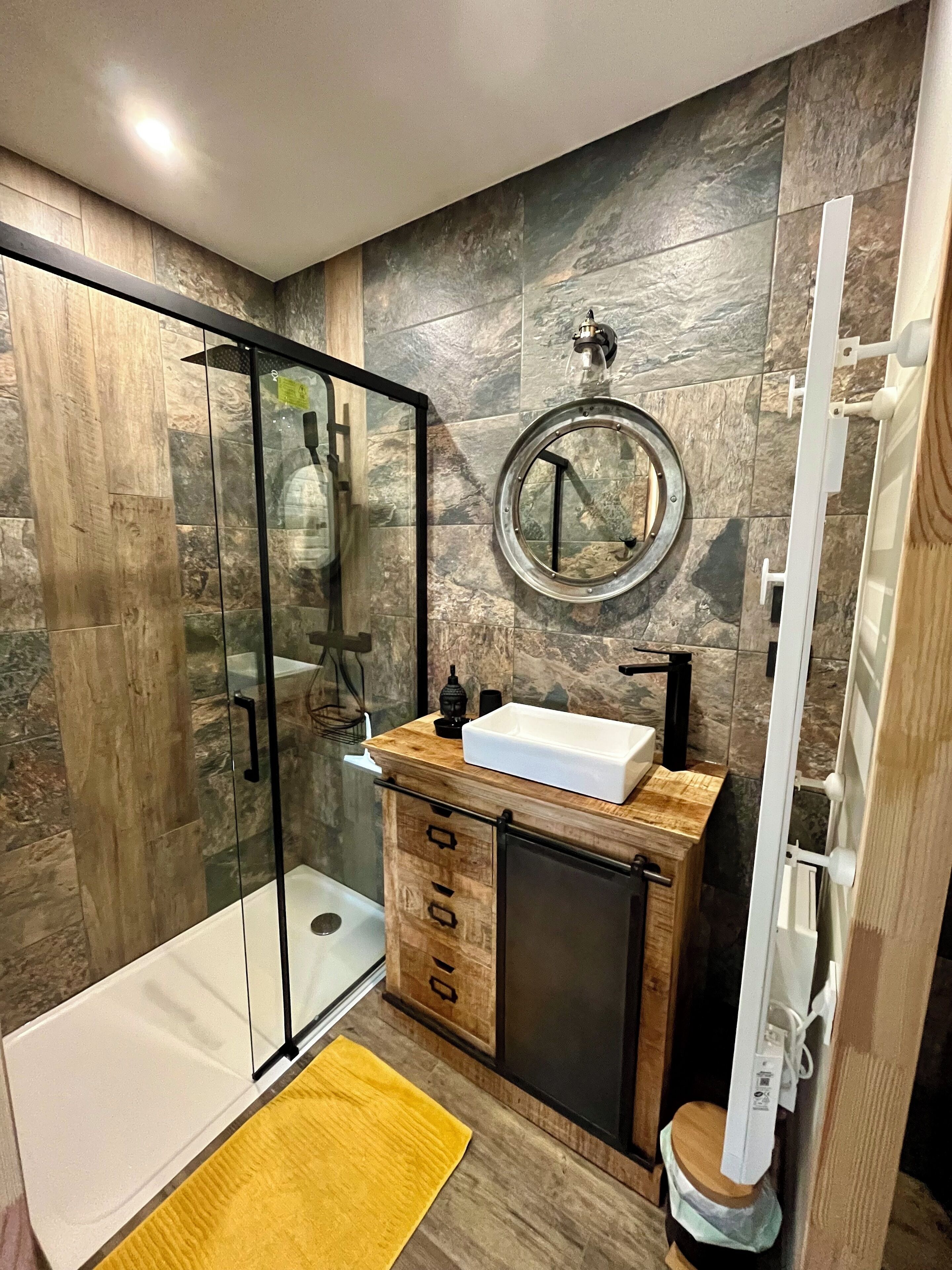 Une salle de bain moderne avec une douche à parois de verre, un meuble-lavabo en bois avec un évier, un miroir et un tapis jaune.
