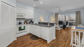 A modern kitchen with white cabinets, a microwave, and a coffee maker. There is a dining area with a table and chairs, and a view of the outdoors through large windows.