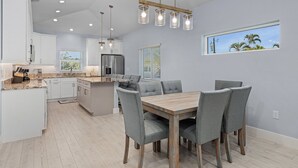 A modern kitchen with white cabinets, a central island, and a dining area with grey chairs and a wooden table. - Beach Time 146 Delmar Ave 3 Bedroom Home by RedAwning (Fort Myers Beach)